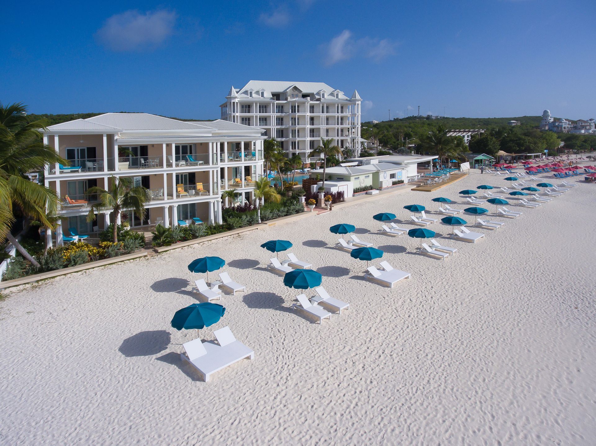 Beach with white sand, turquoise umbrellas, and resort buildings under a blue sky.