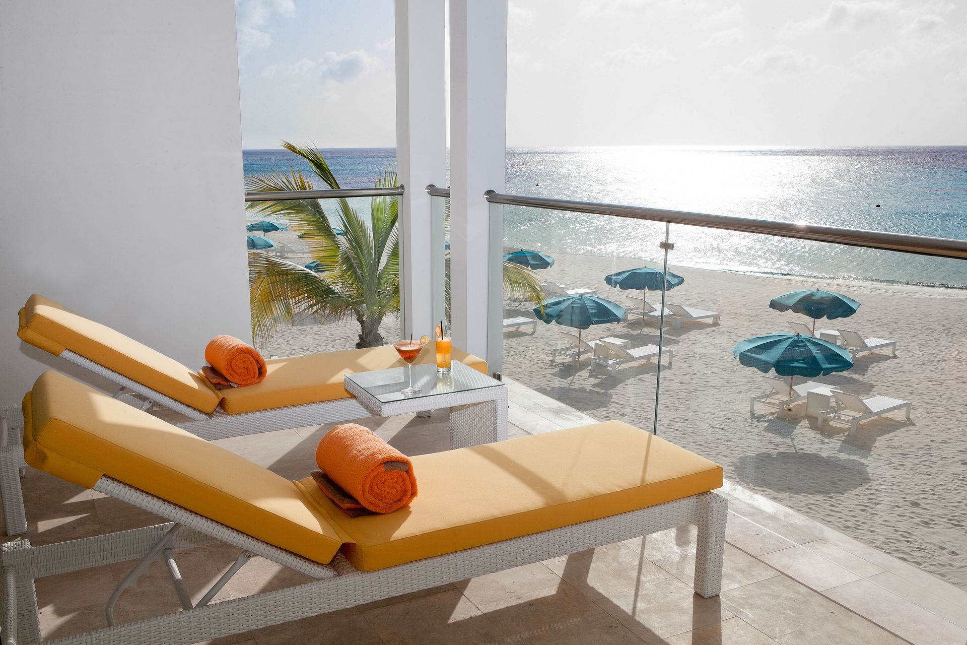 Balcony with beach view: yellow lounge chairs, cocktails, ocean, umbrellas.