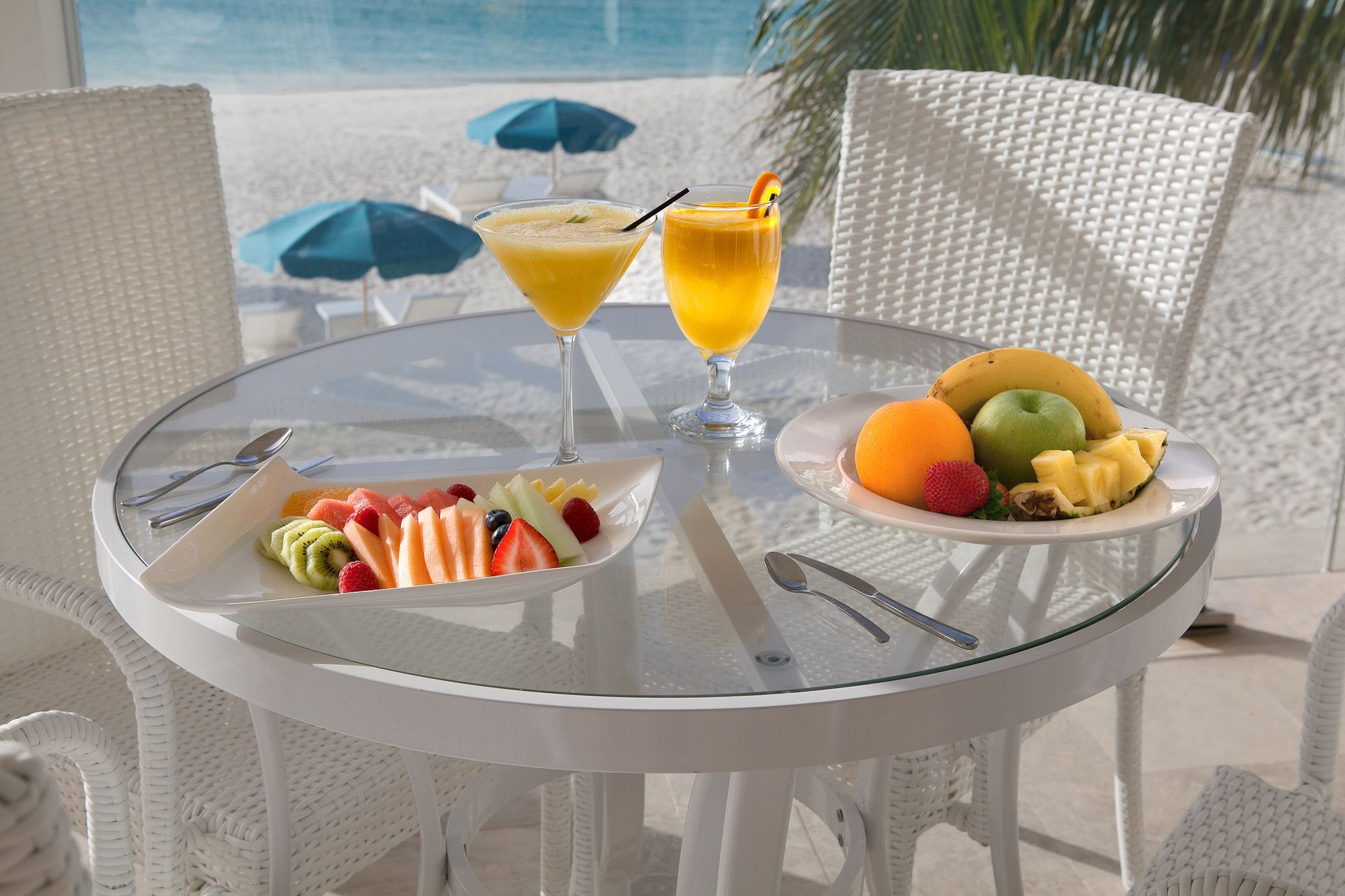 Round table with fruit and cocktails, white wicker chairs, beach view with blue umbrellas.