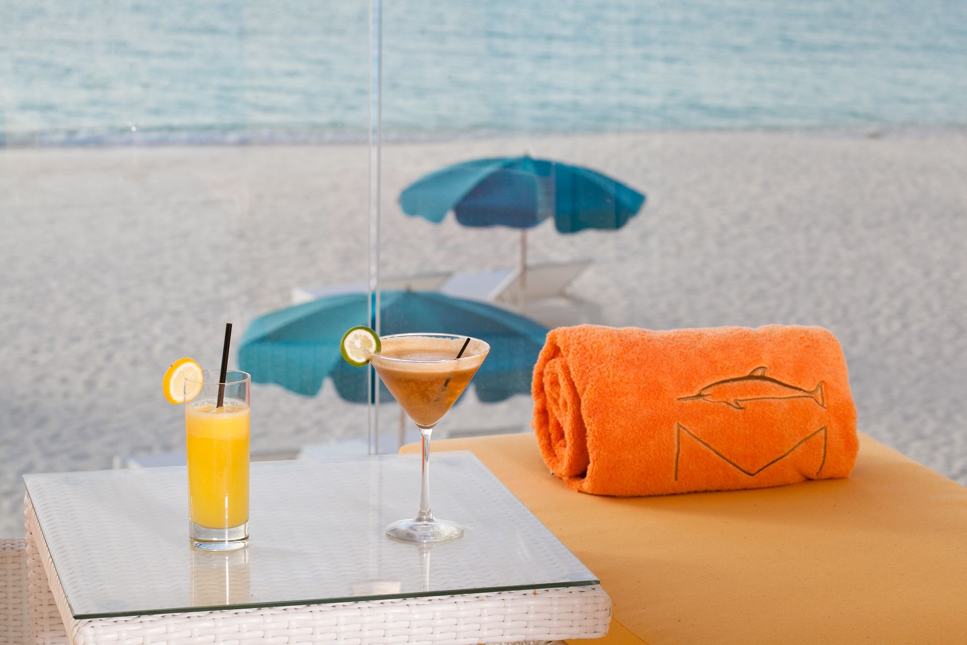 Cocktails on a table with a beach view: orange towel, turquoise umbrellas, ocean, and sand.