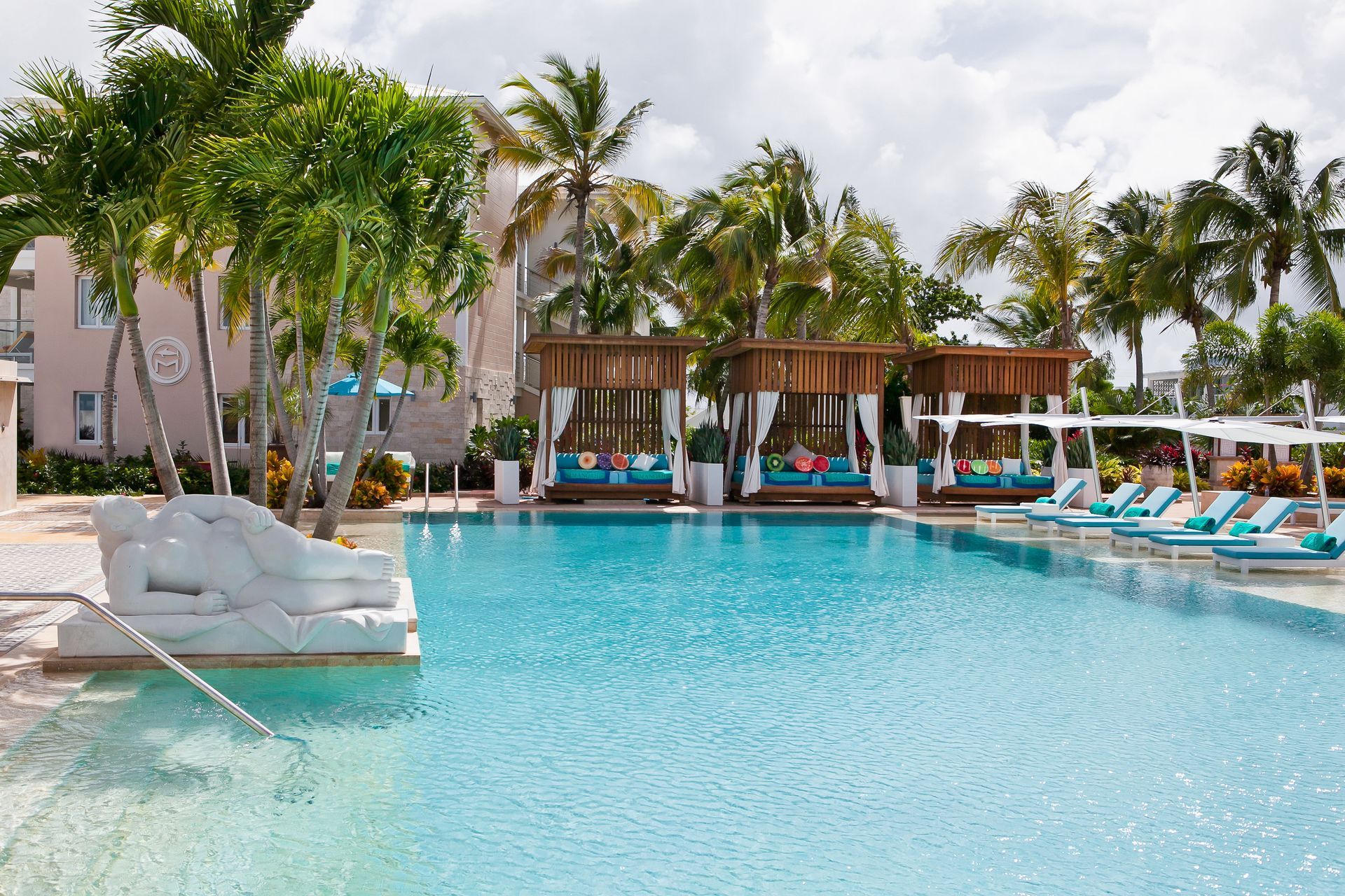 Swimming pool with cabanas and lounge chairs at a resort, palm trees, and a bright sky.