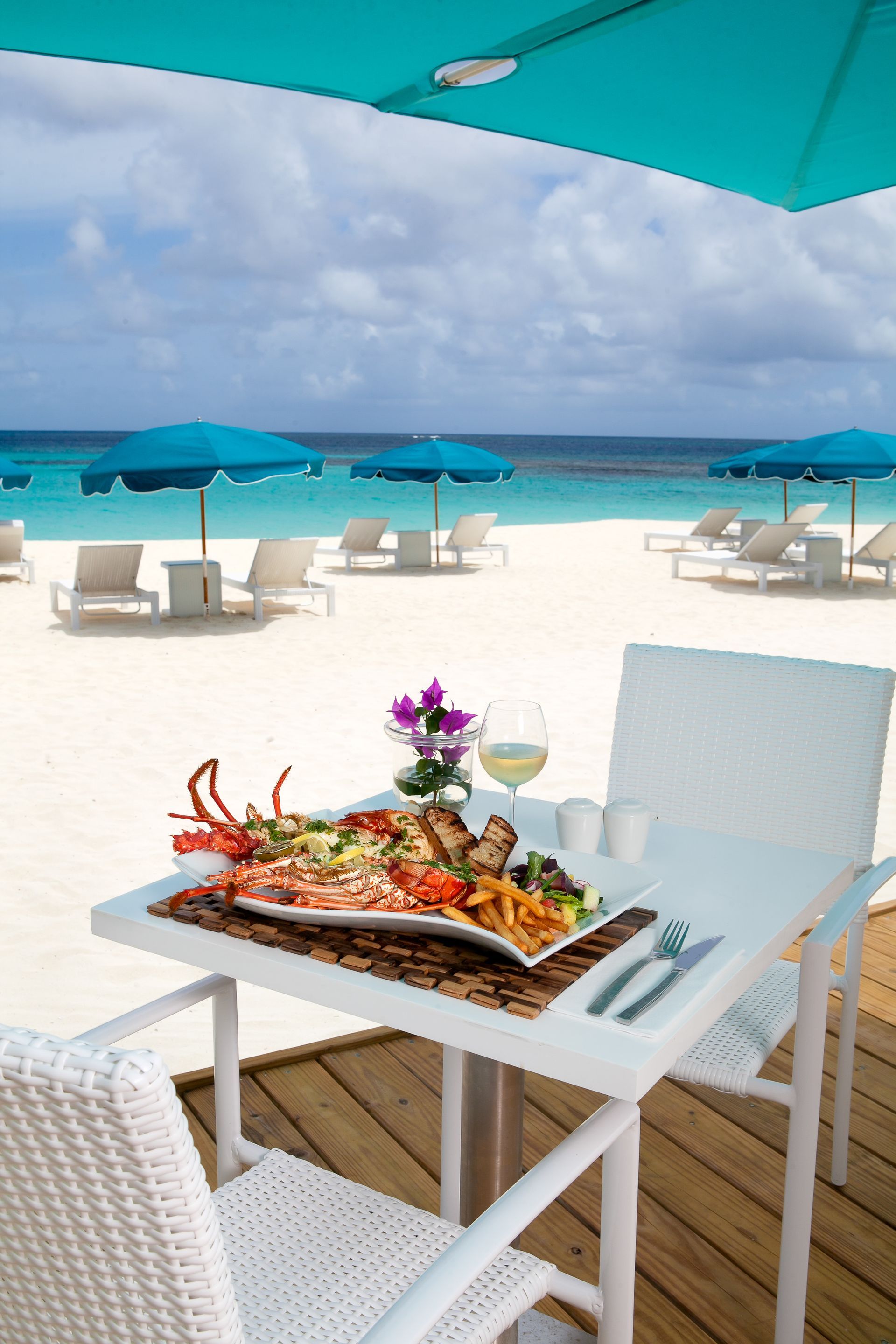 Table set for lunch on a beach with a seafood platter, white chairs, blue umbrellas, and ocean.