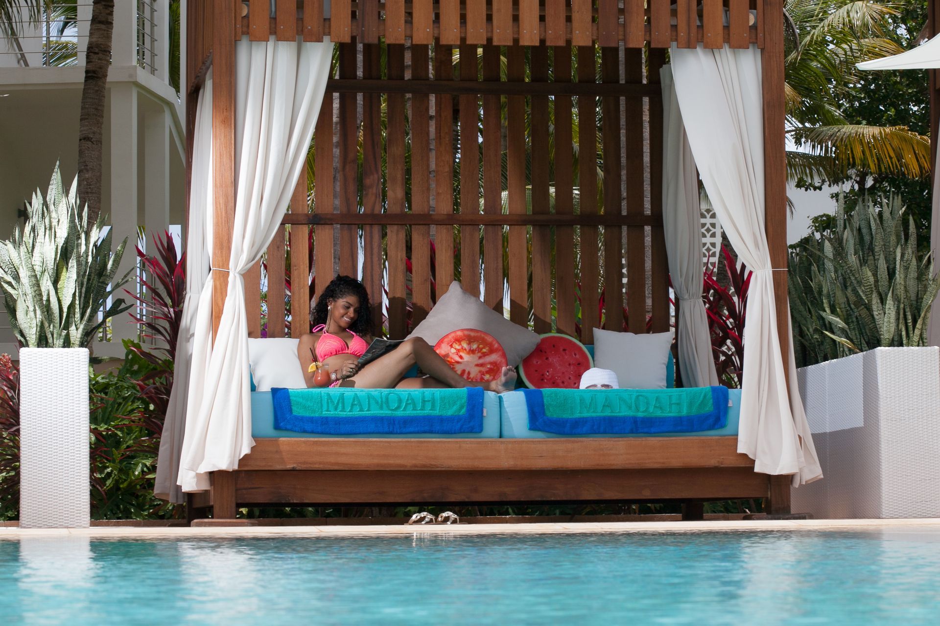 Woman relaxing on a shaded lounge bed by a pool, wearing pink swimwear, looking at a device.