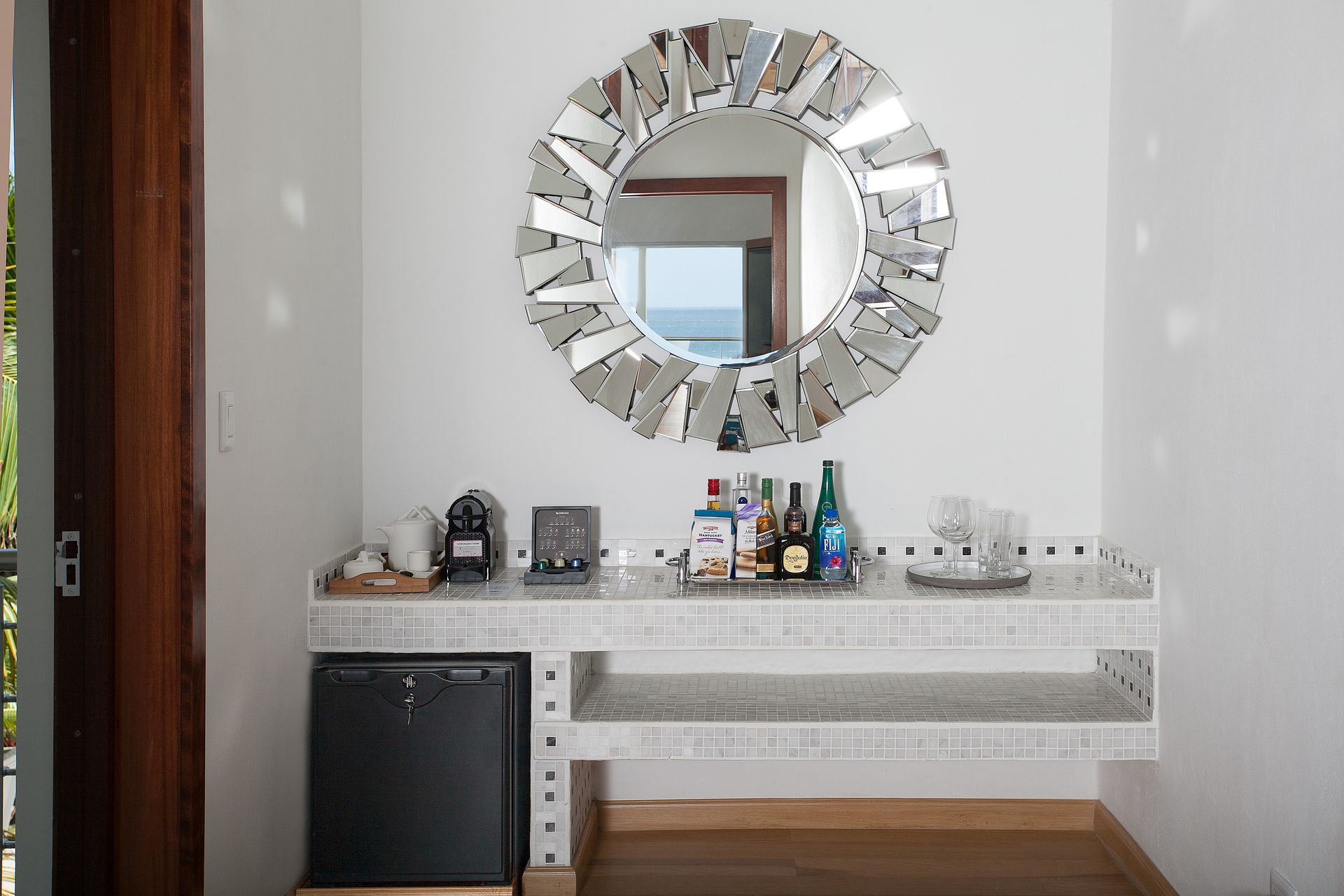 White bar area with a decorative mirror, drinks, and a small fridge.