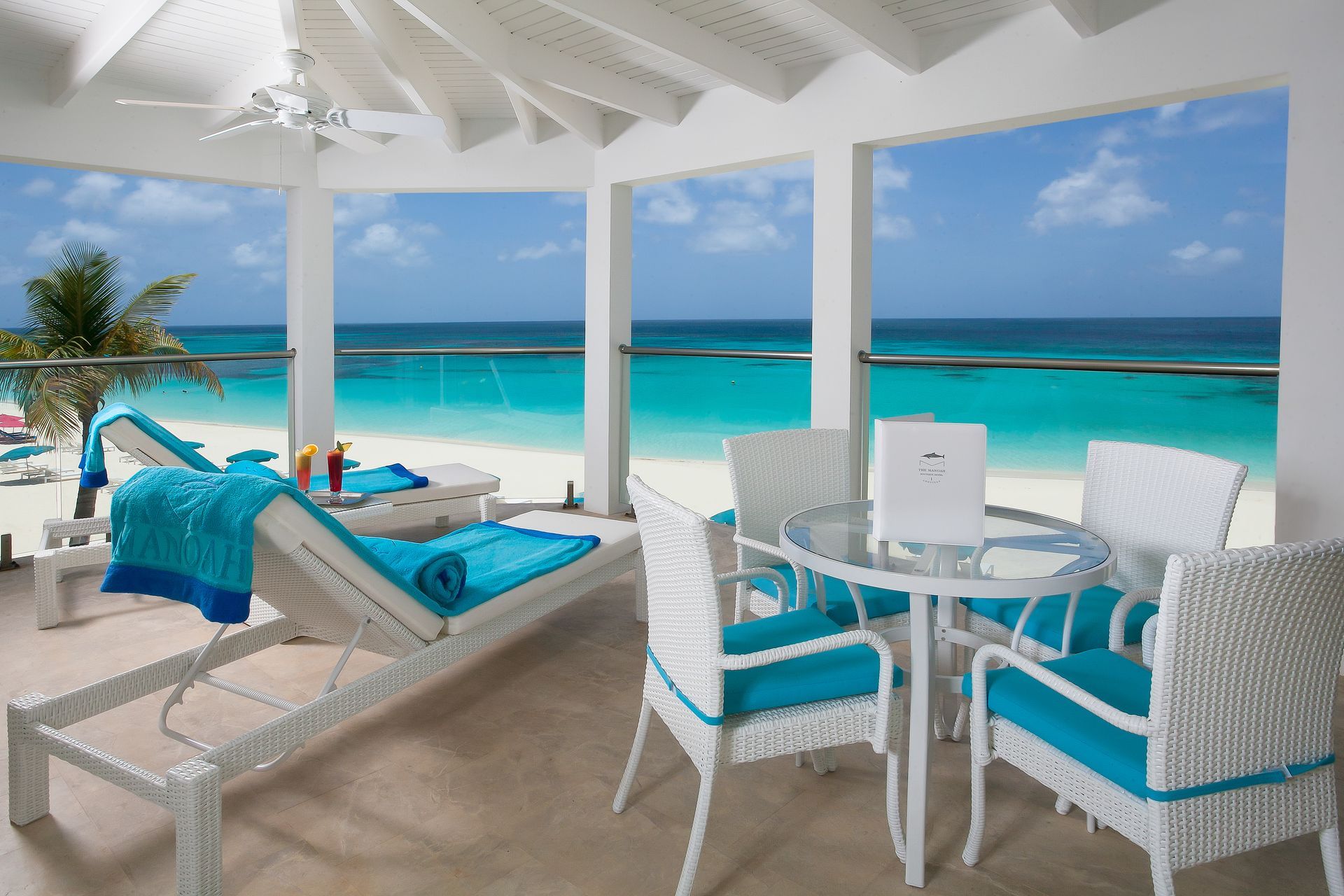 Beachfront patio with white furniture, turquoise cushions and towels, overlooking the ocean.
