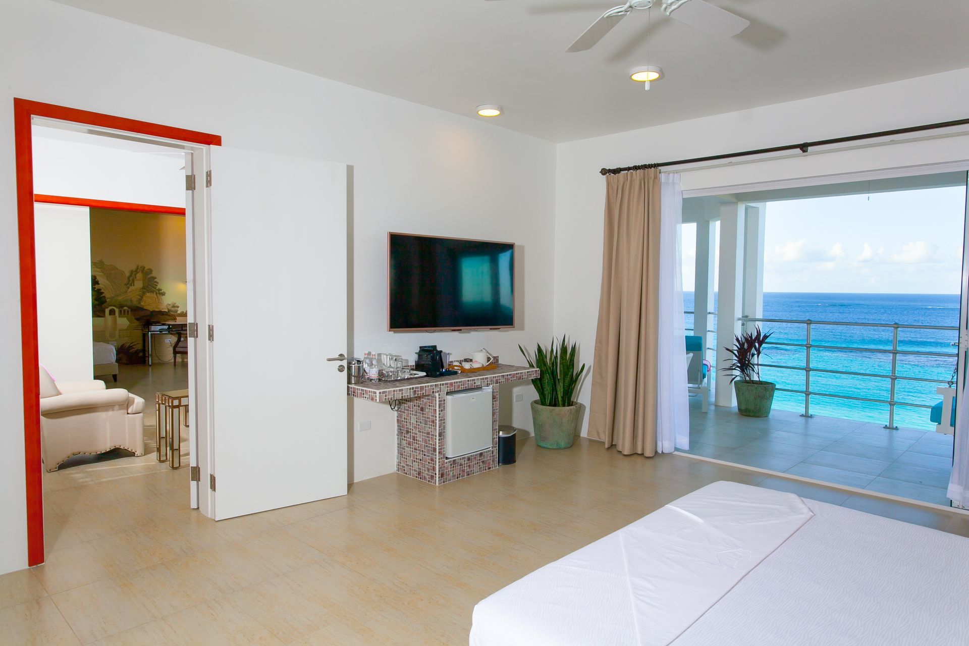Bright bedroom with ocean view balcony, TV, and open door to a lounge area.
