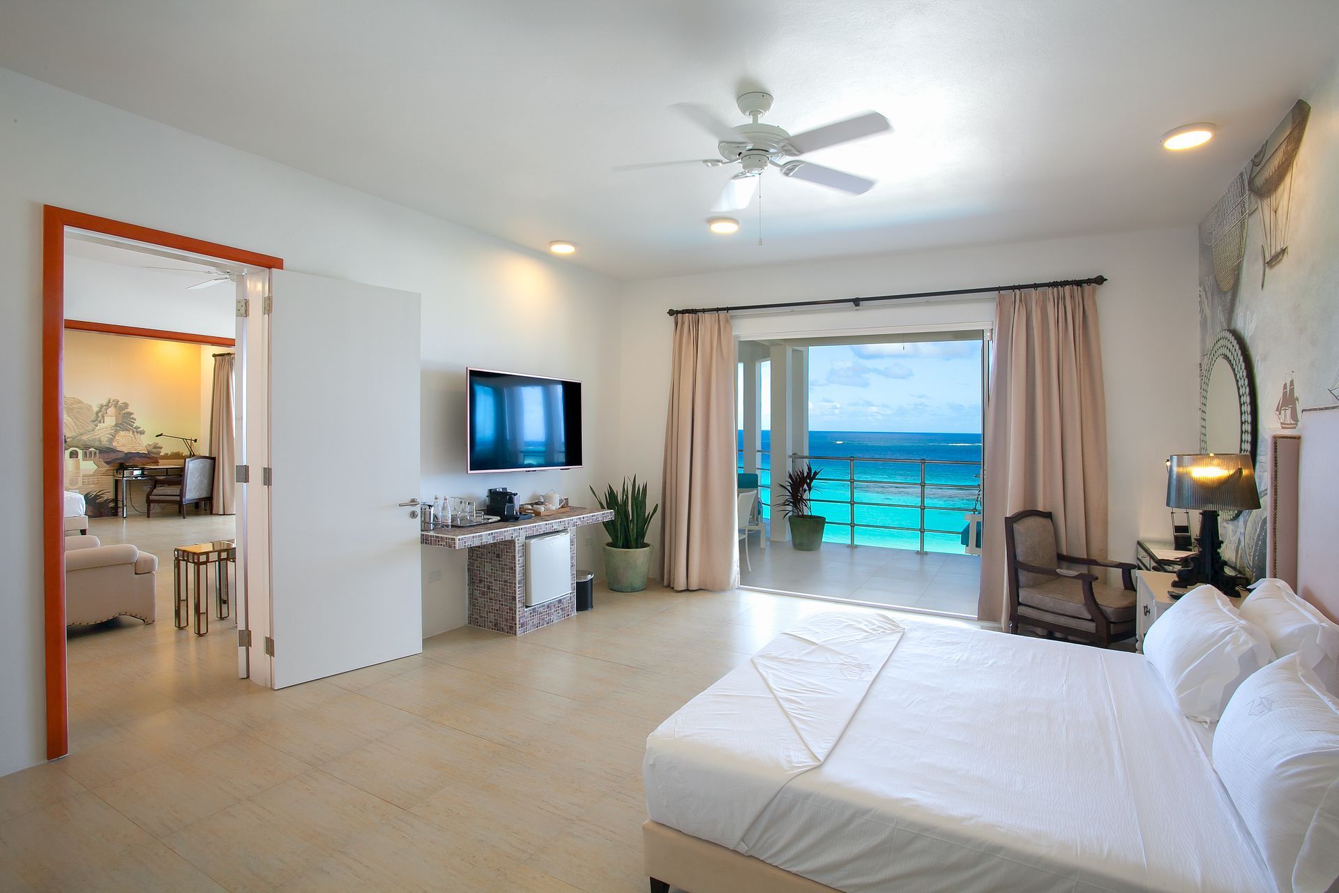Bedroom with ocean view, white walls, balcony, bed, TV, and a glimpse of a dining area.