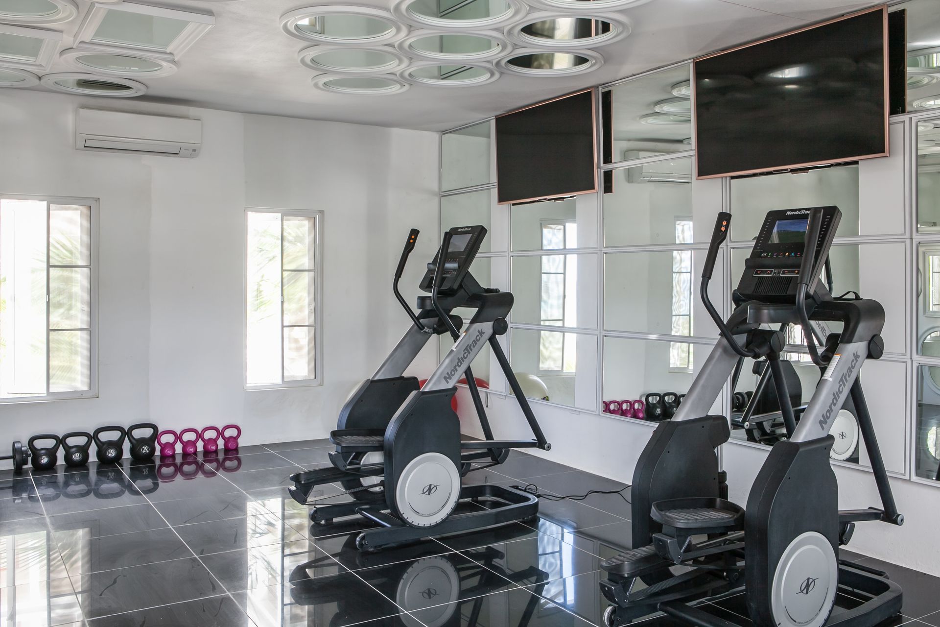 Gym with elliptical machines, weights, mirrors, and TVs in a bright, white room.