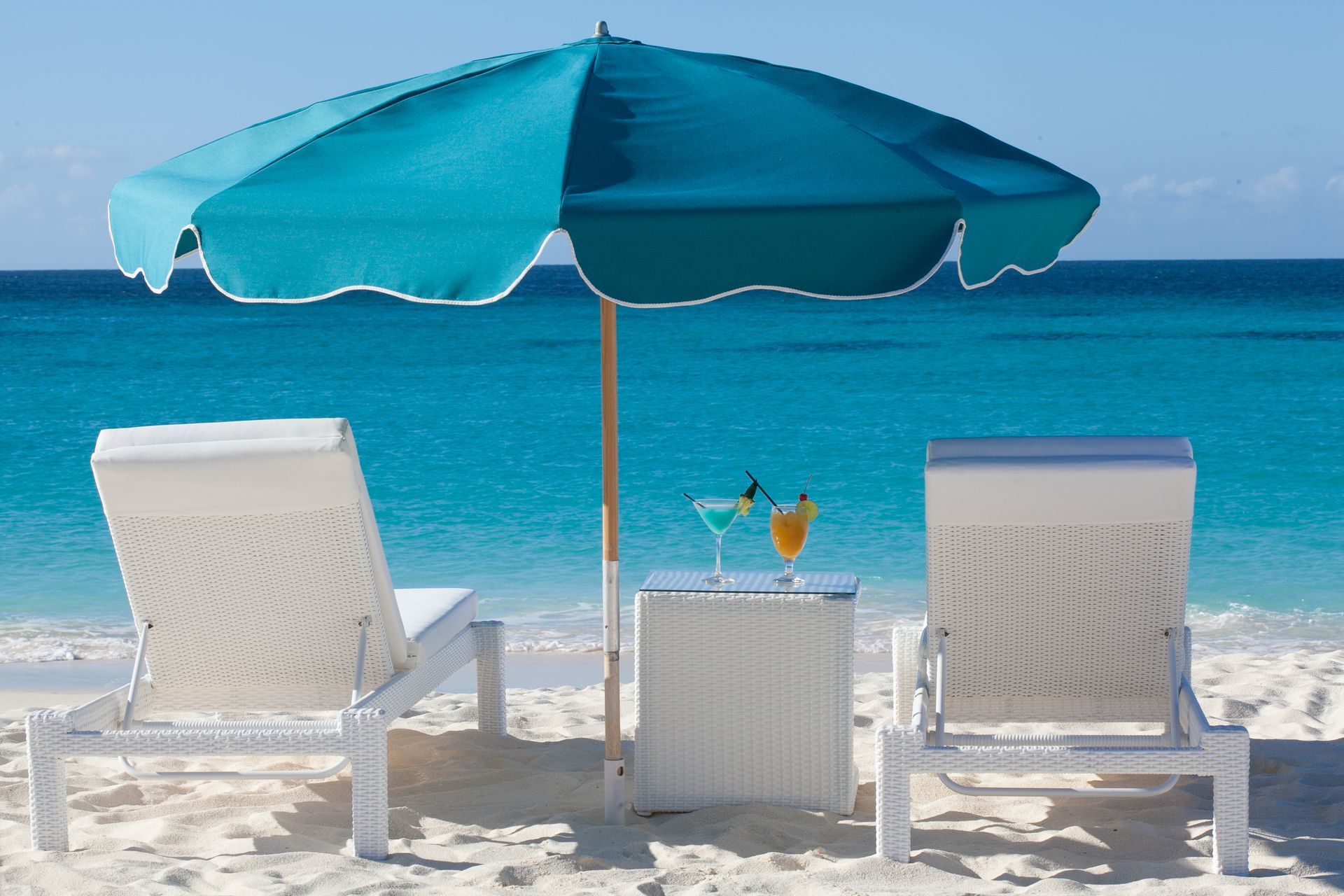 Two white lounge chairs, a turquoise umbrella, and cocktails on a table, on a sandy beach.