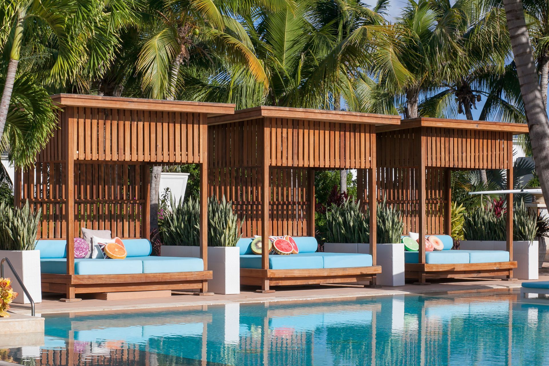 Poolside cabanas with blue cushions, wood frames, tropical setting.