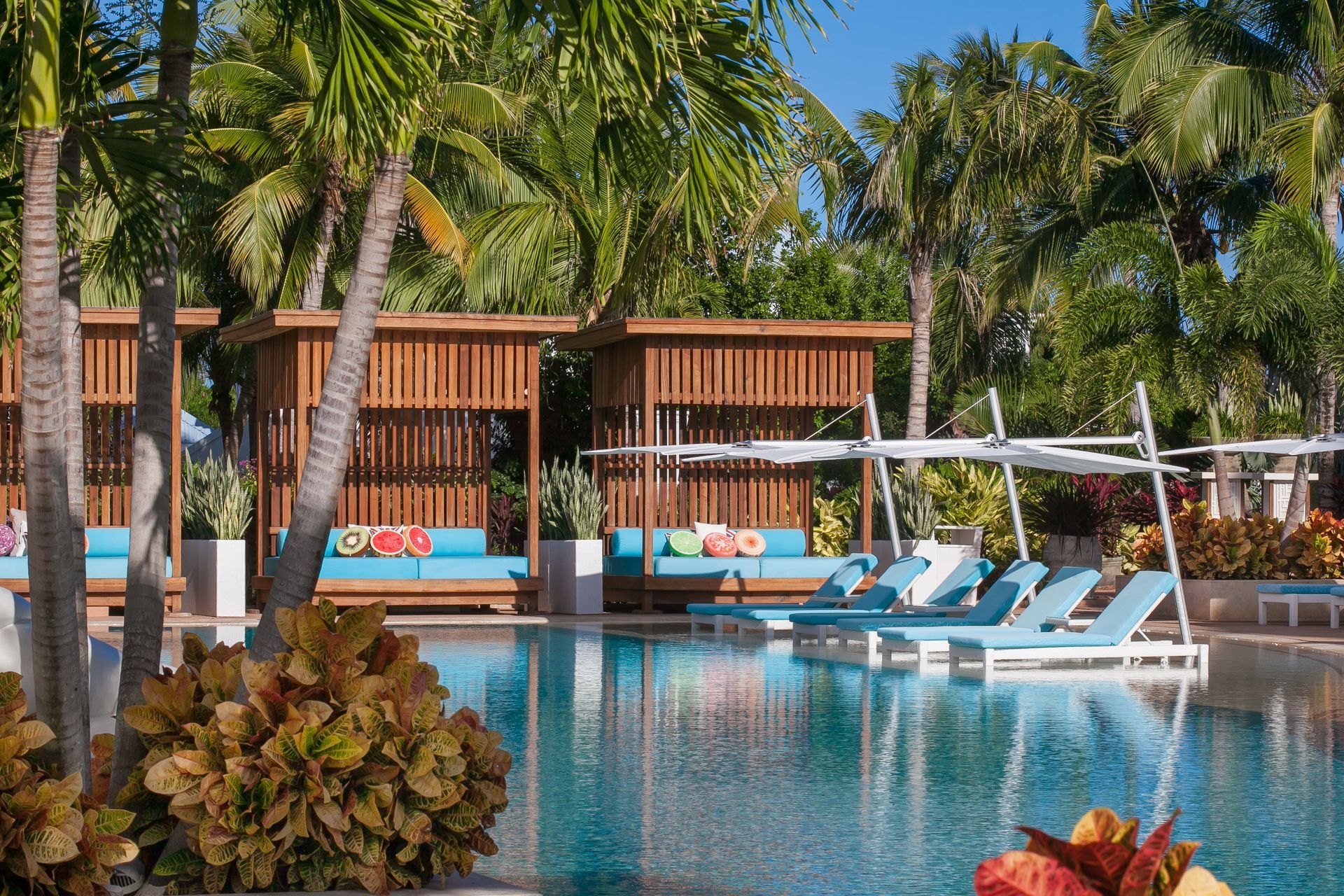 Poolside cabanas with blue cushions, sun loungers, palm trees, and clear blue water.