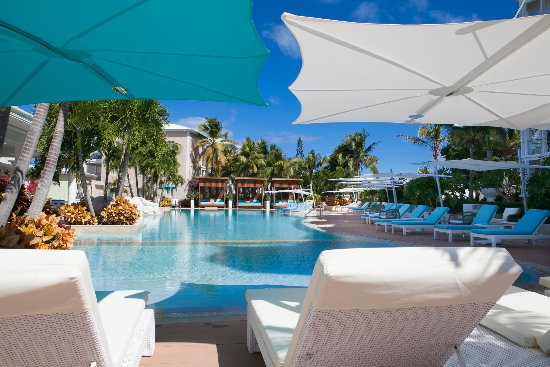 Poolside scene: white lounge chairs, turquoise pool, blue sky, white and teal umbrellas, resort setting.