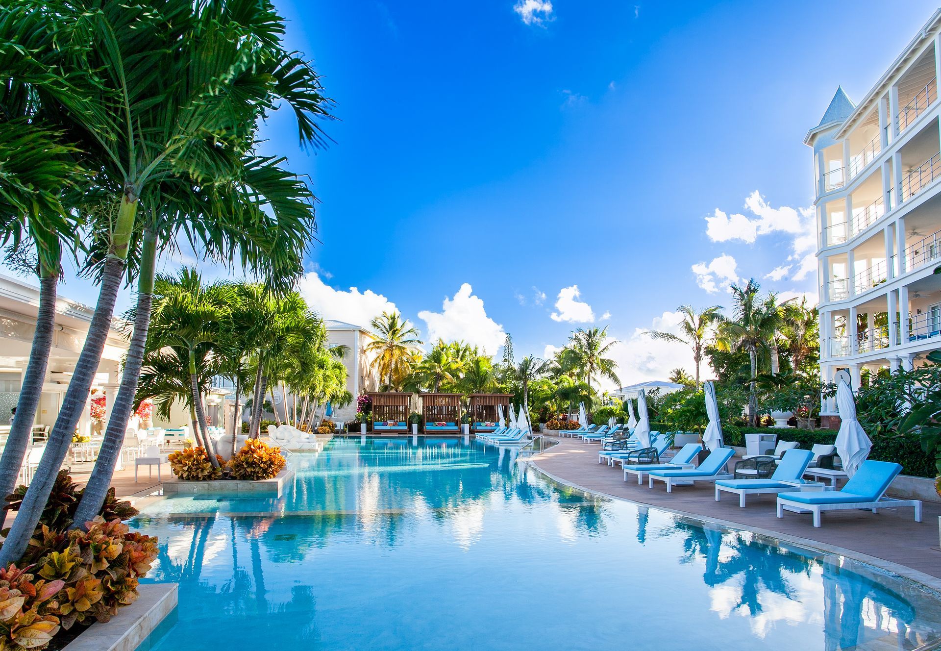 Pool with lounge chairs, palm trees, and resort buildings against a bright blue sky.