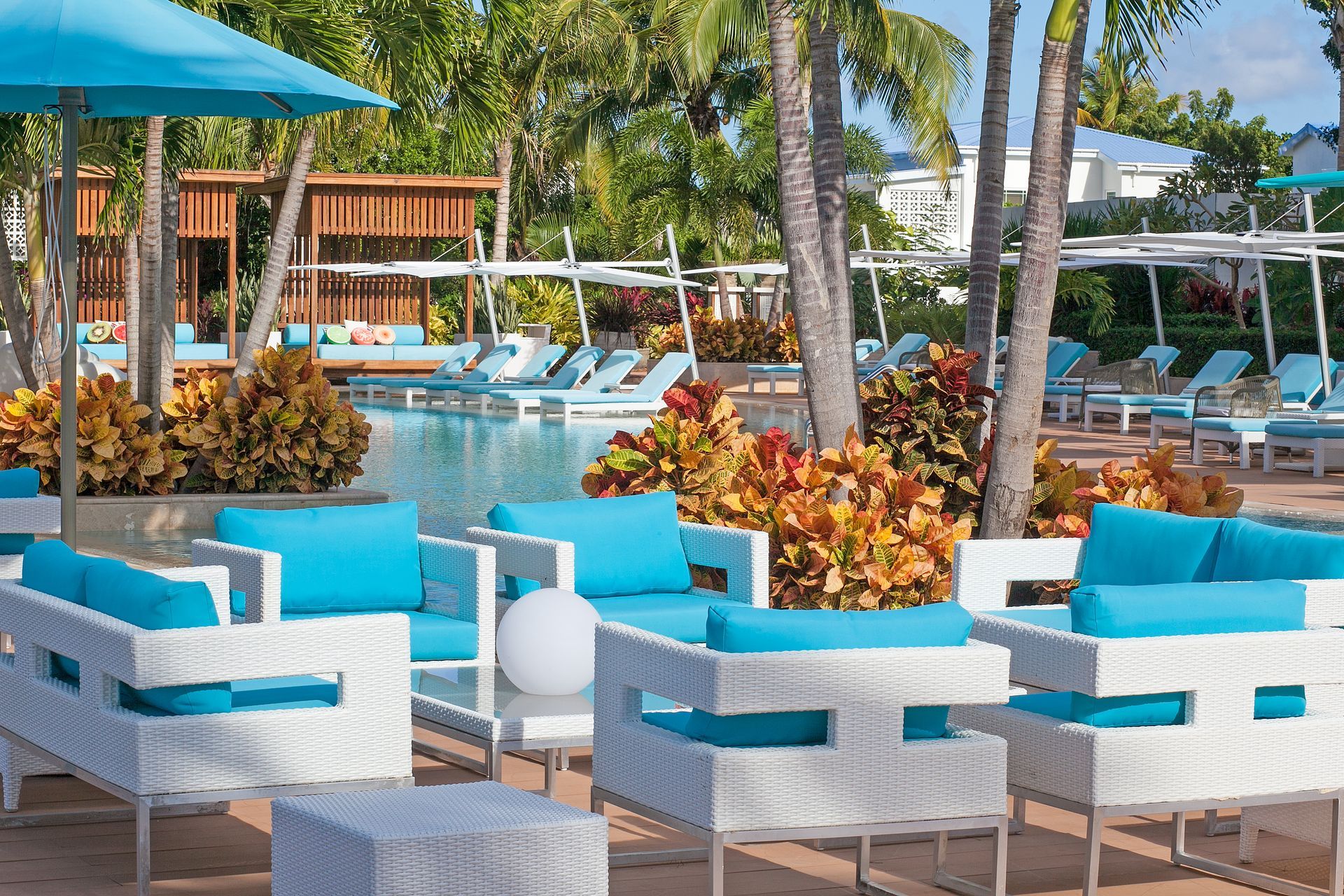 Poolside lounge area with white furniture, turquoise cushions, and parasols. Lush tropical plants.