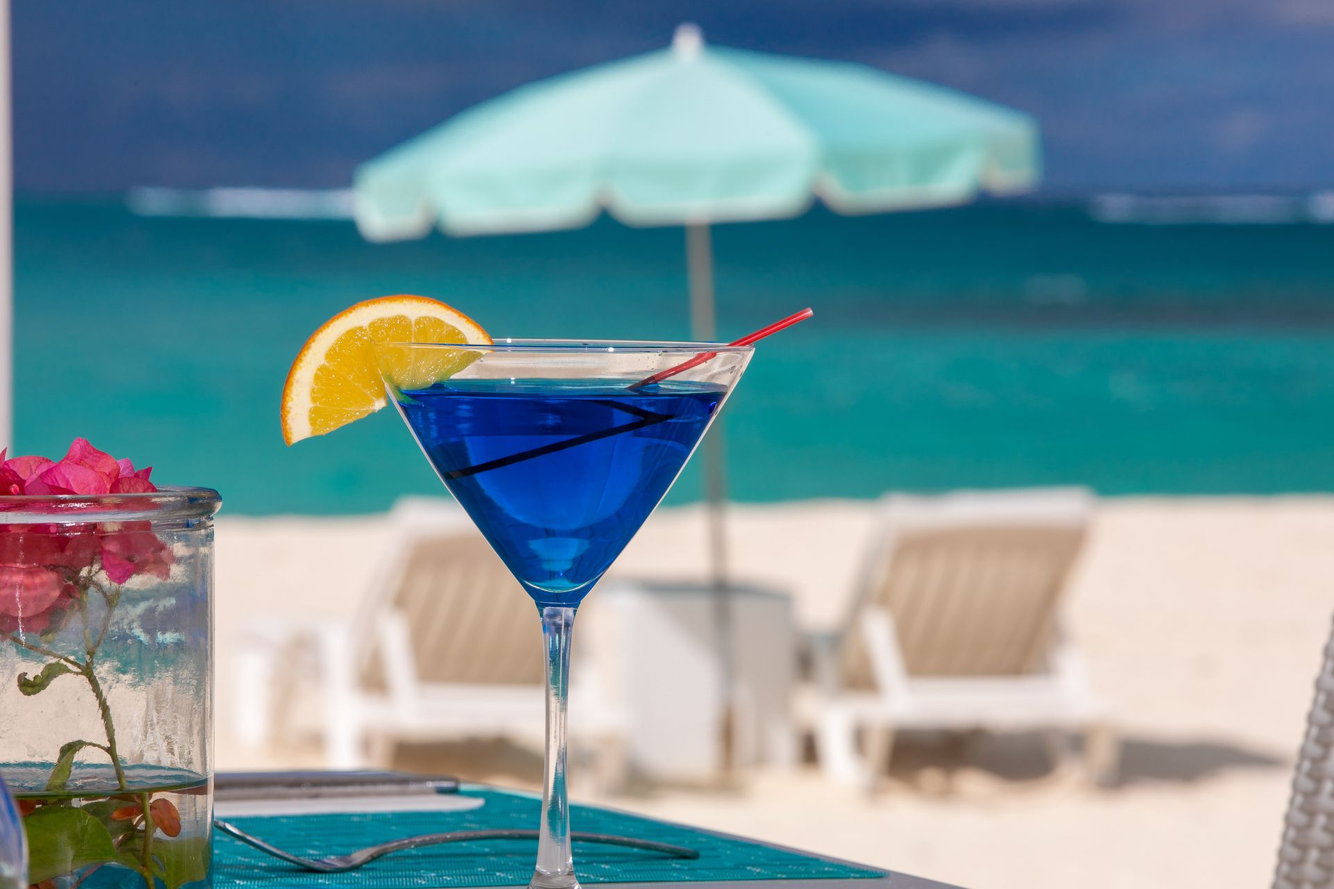 Blue cocktail on a beach table with orange slice garnish, beach, parasol, and chairs in background.