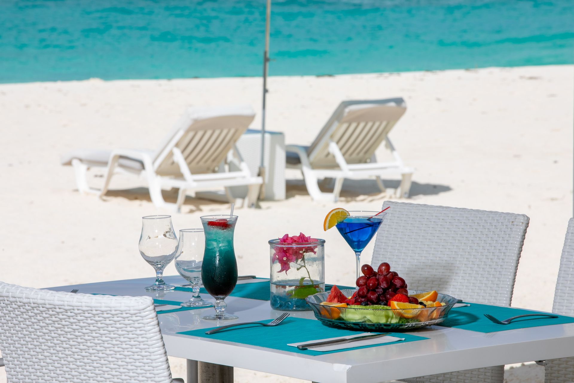 Table set for beach dining: cocktails, fruit, white chairs, turquoise placemats, ocean view.