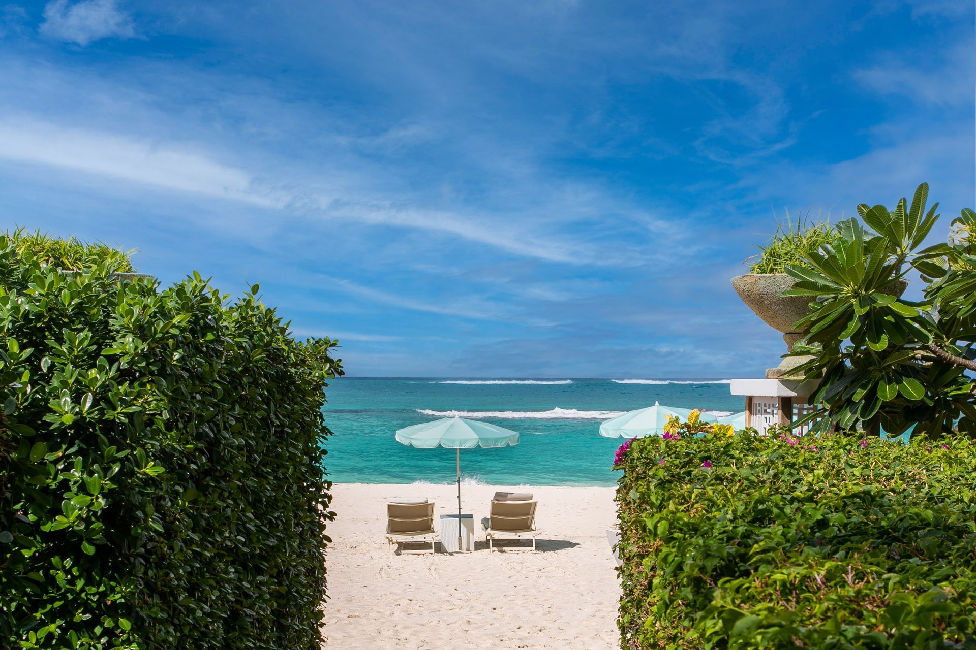 Beach access with two lounge chairs and umbrellas, framed by green bushes, against a blue sky.