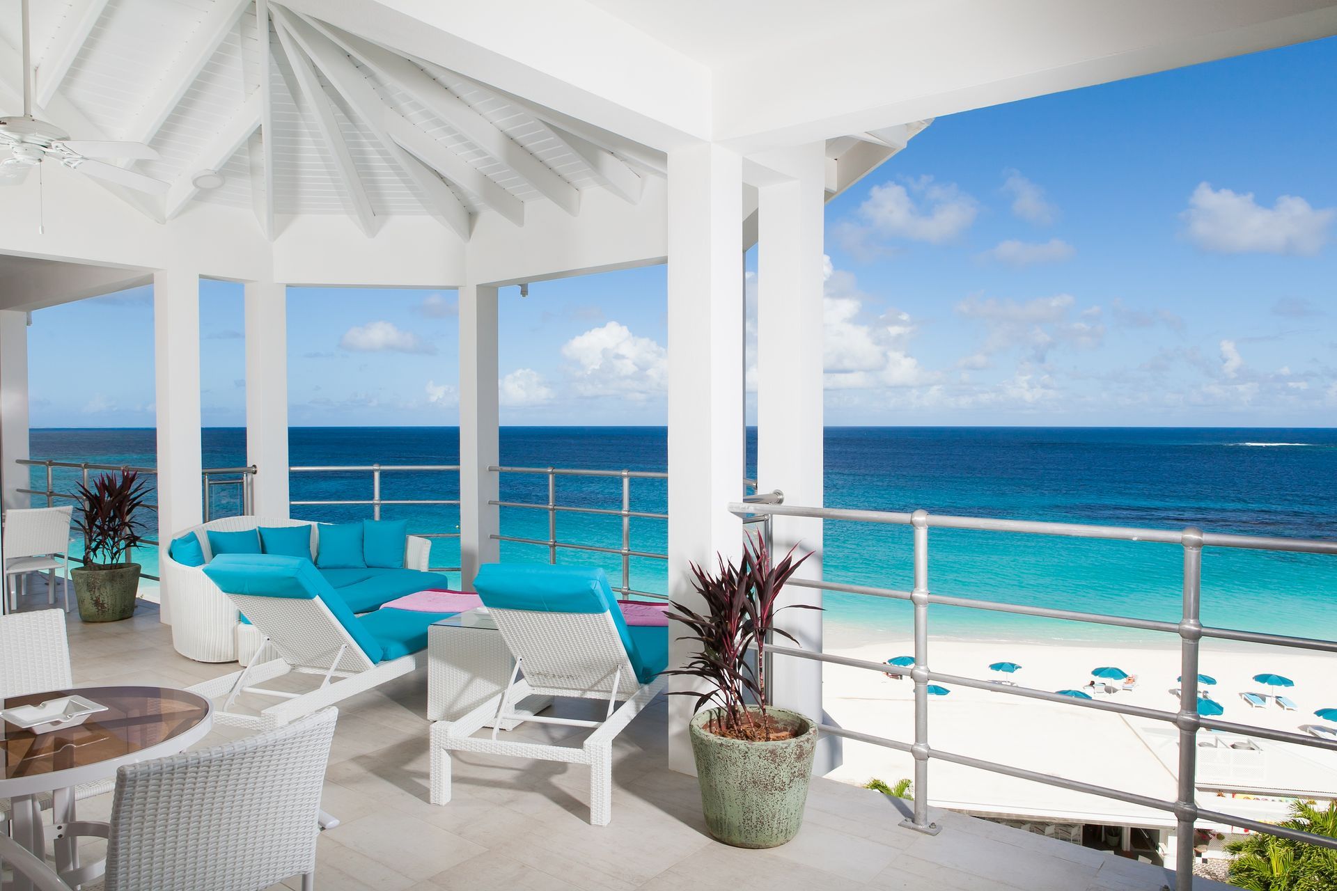 Oceanfront balcony with white and teal furniture, overlooking a turquoise sea and sandy beach.