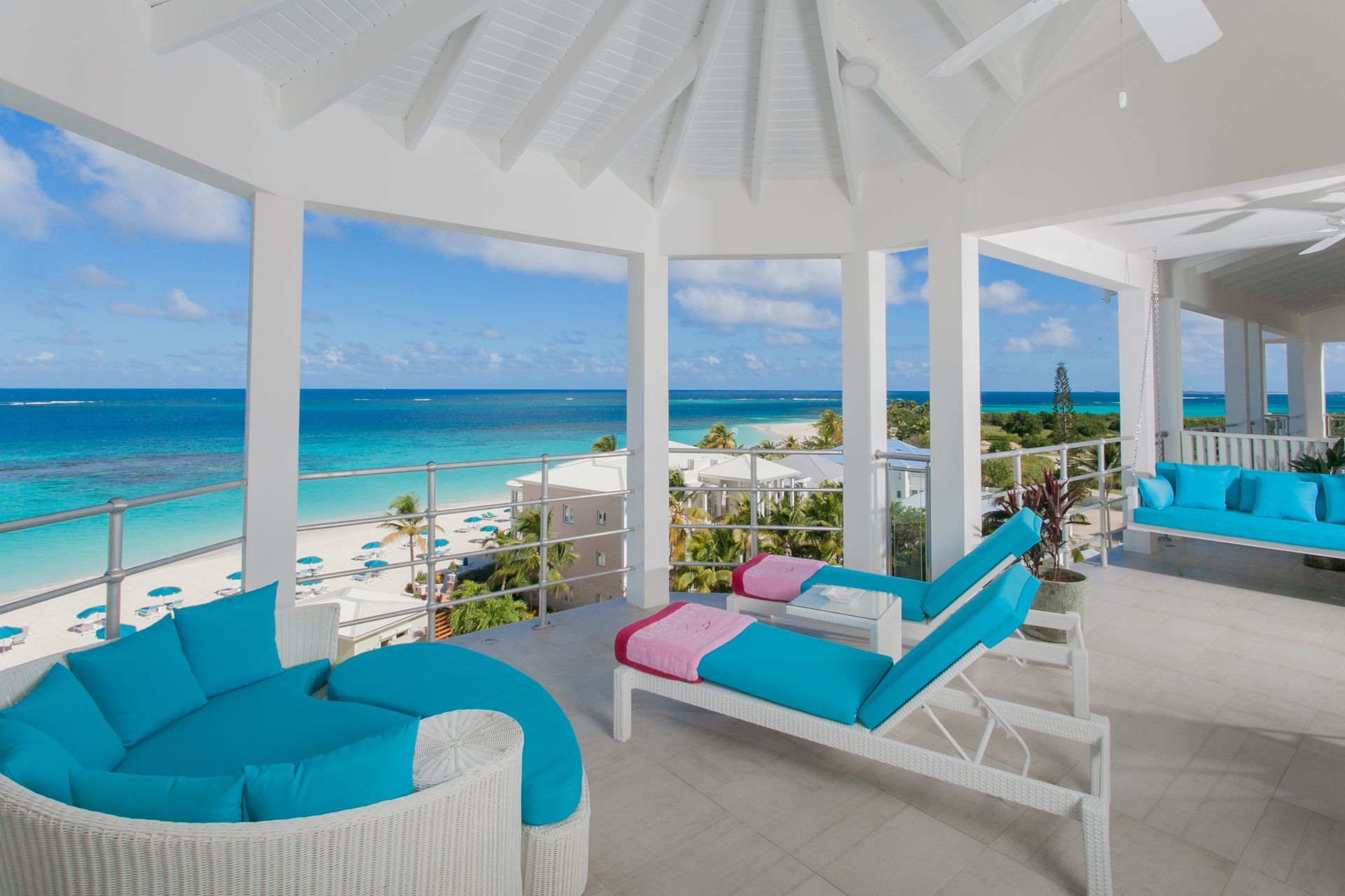 Bright white gazebo overlooking a turquoise ocean and beach with blue seating and colorful cushions.