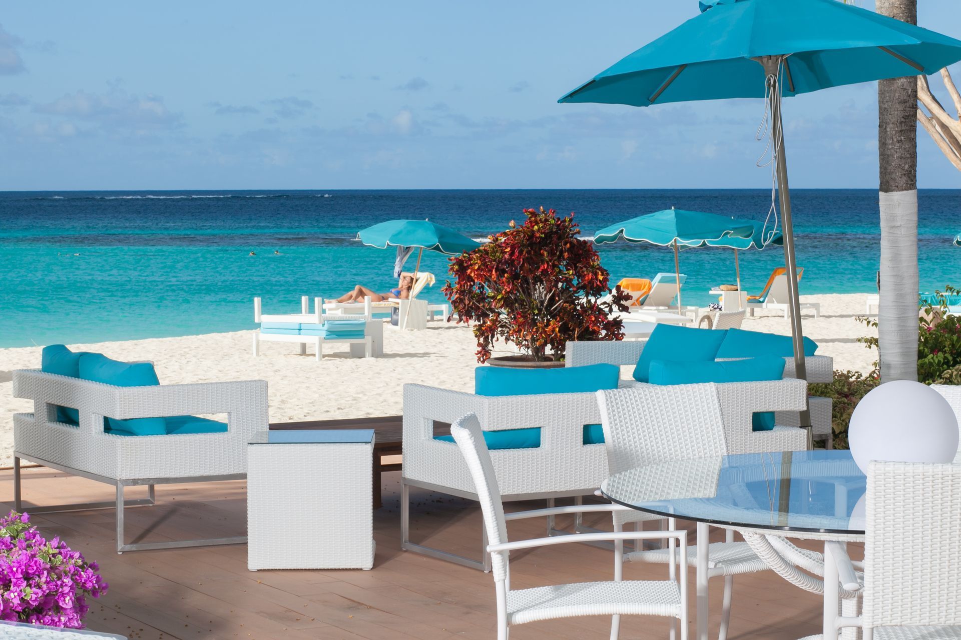 Beachside lounge area with white furniture, turquoise cushions, umbrellas, and ocean view.