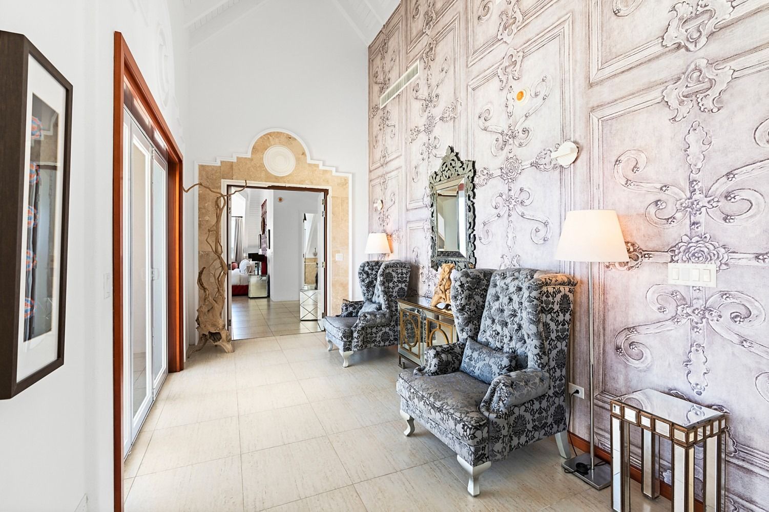 Elegant hallway with baroque wallpaper, two patterned armchairs, mirror, and a standing lamp.
