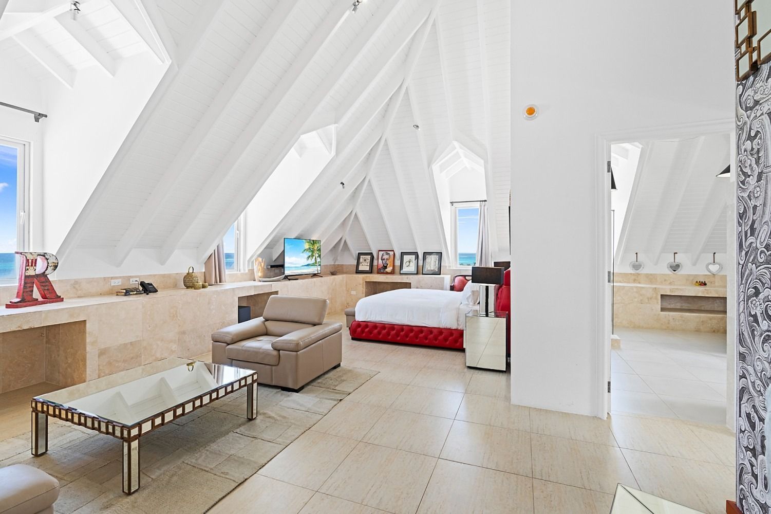 Spacious white interior with a bed, couch, TV, and ocean views, illuminated by sunlight.