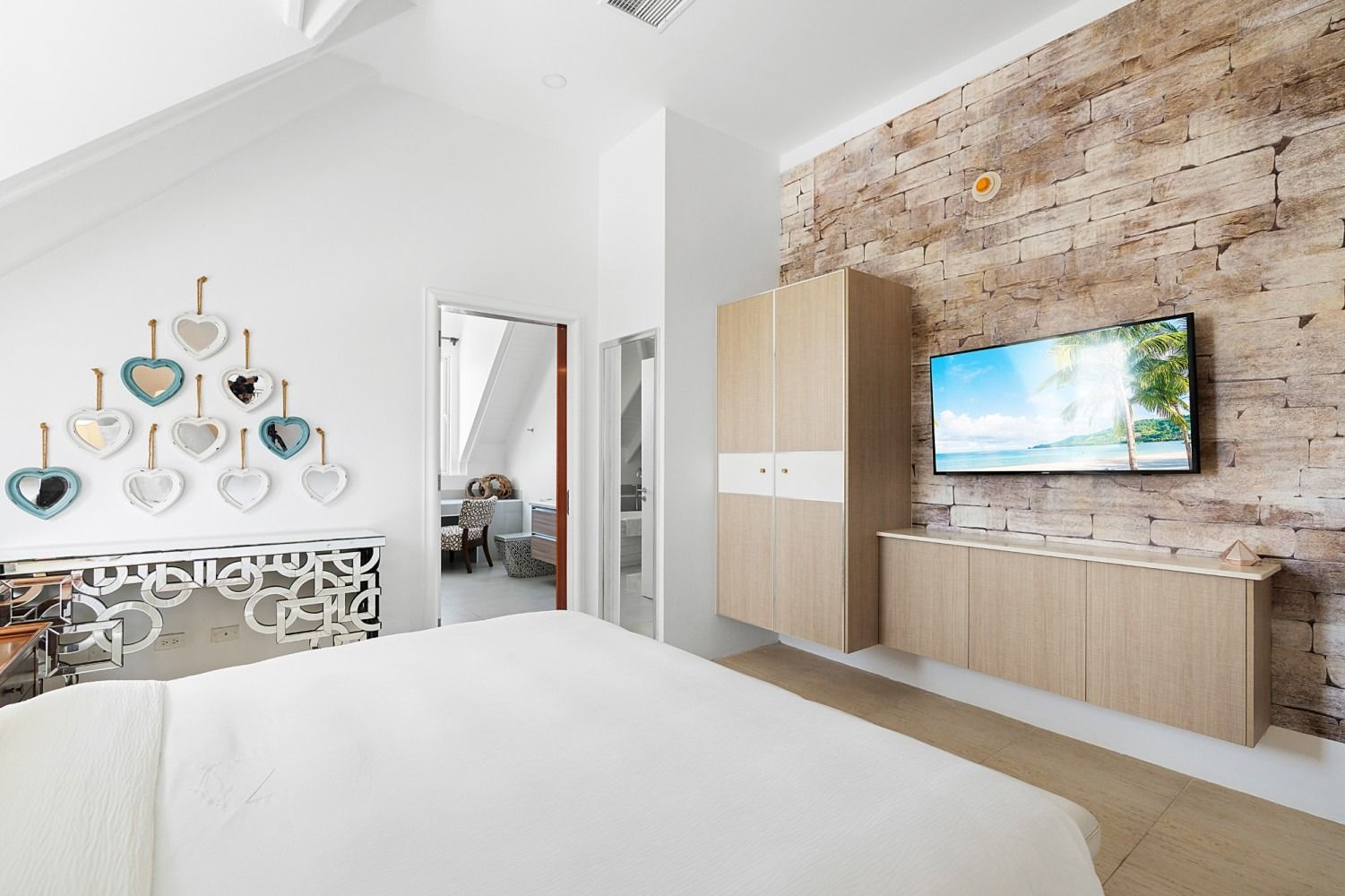 Bedroom with white walls, brick accent wall, bed, TV, wooden cabinets, and decorative hearts.