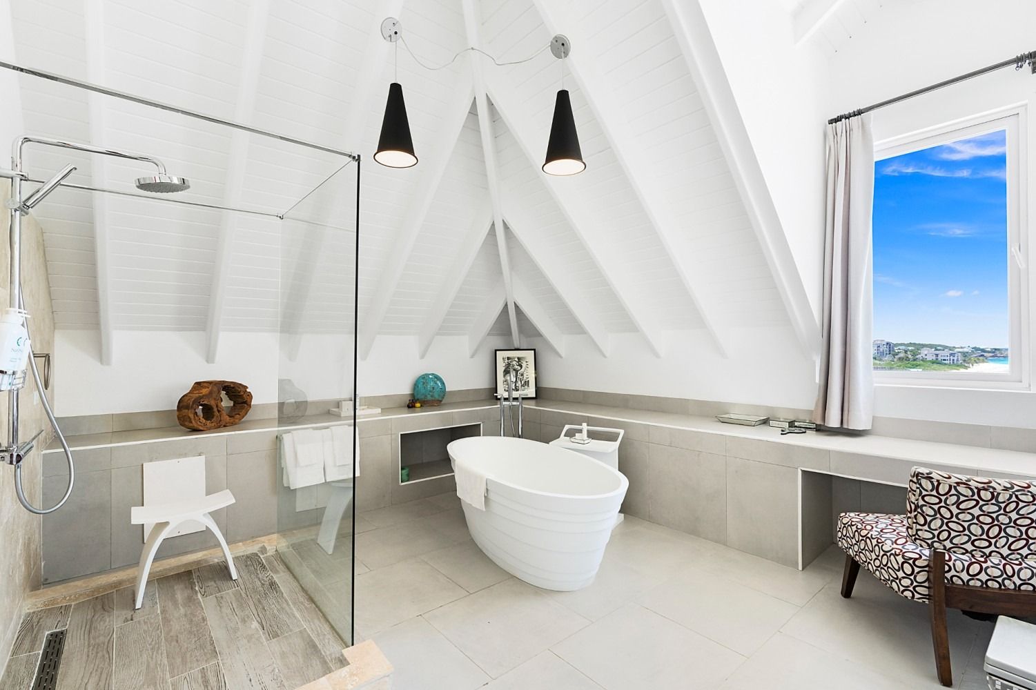 Bright, airy bathroom with glass shower, freestanding tub, window with ocean view, and decorative lights.