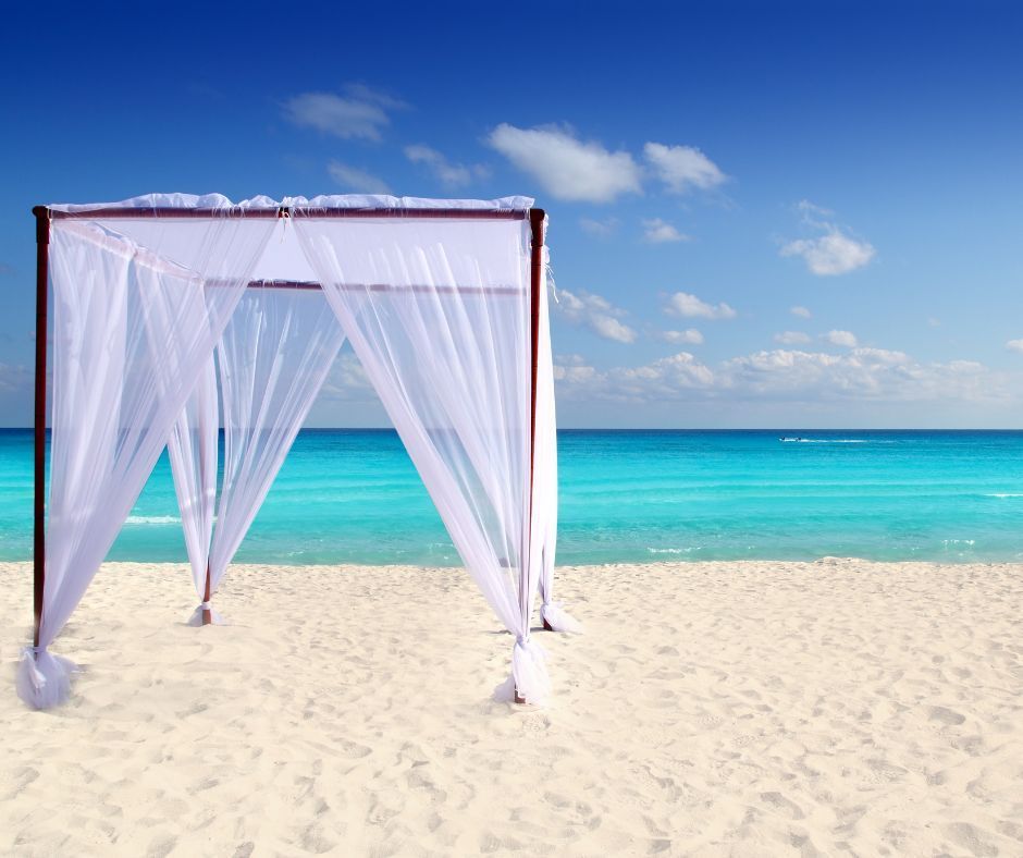 Beach canopy with white sheer curtains on a sandy beach with turquoise water and blue sky.