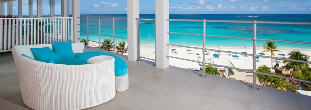 A beach scene with a white cushioned sofa, turquoise pillows, and a view of the ocean from a balcony.