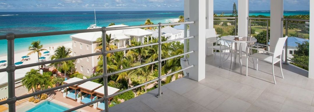 Balcony overlooking a turquoise ocean, sandy beach, and lush greenery. White resort.