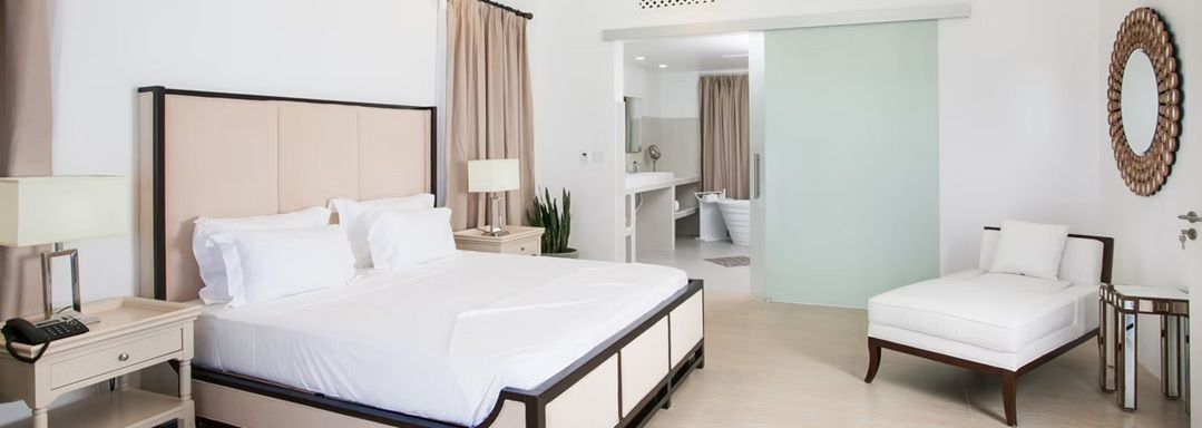 Bedroom with a large bed, chaise lounge, and a sliding door leading to a bathroom. Neutral colors.