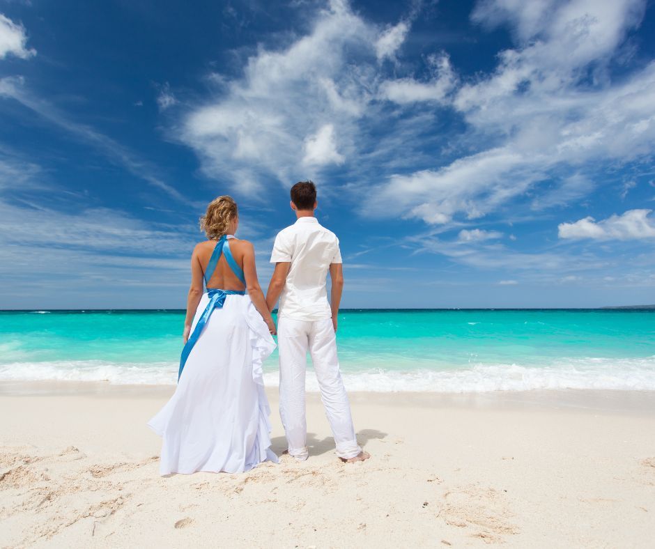 Couple in wedding attire standing on a beach, looking at turquoise water under a blue sky.
