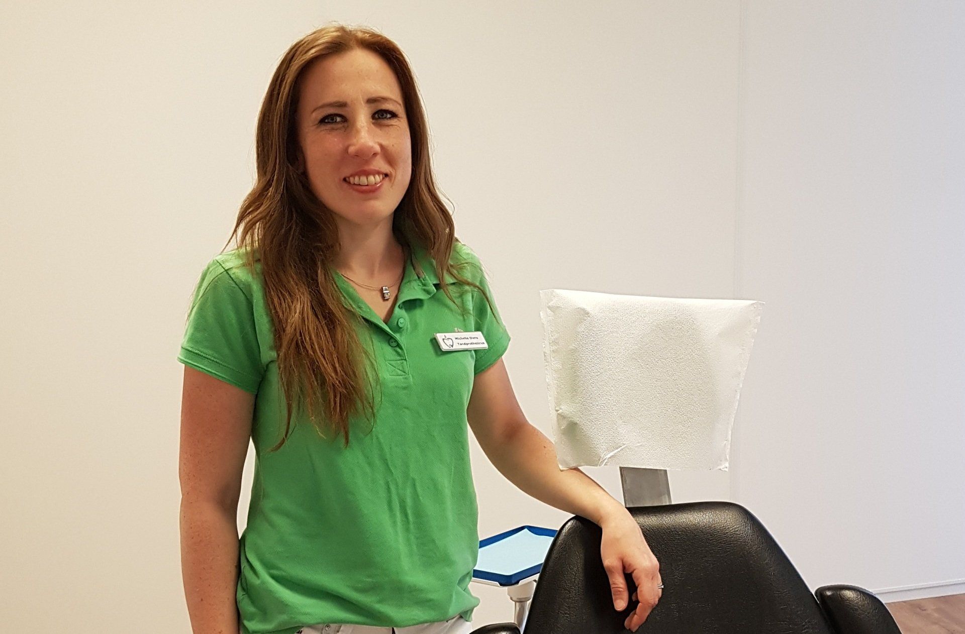 Een persoon in een groen poloshirt met naambadge staat naast een zwarte tandartsstoel in een lichte, minimalistische ruimte.