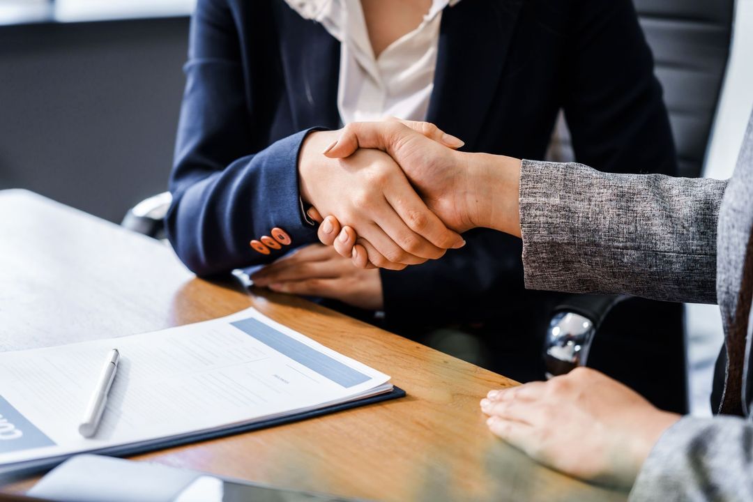 Two people in business attire shaking hands across a meeting table with papers and a notebook nearby