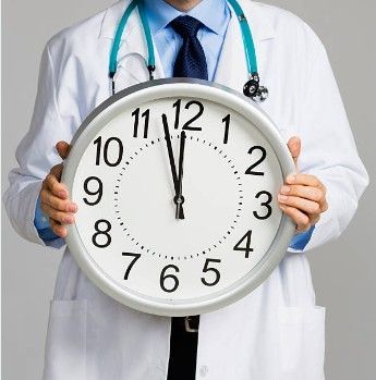 Doctor holding a clock, hands at 12:00. He wears a white coat and stethoscope.