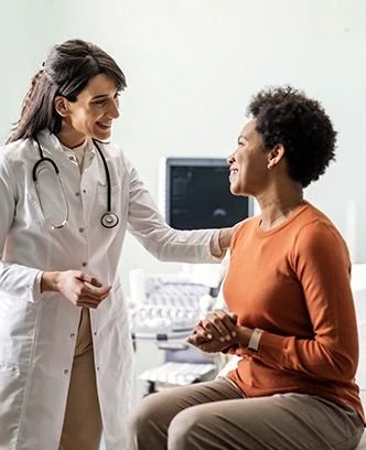 A doctor in a white coat comforts a patient in an orange shirt. Hospital setting, both smiling.