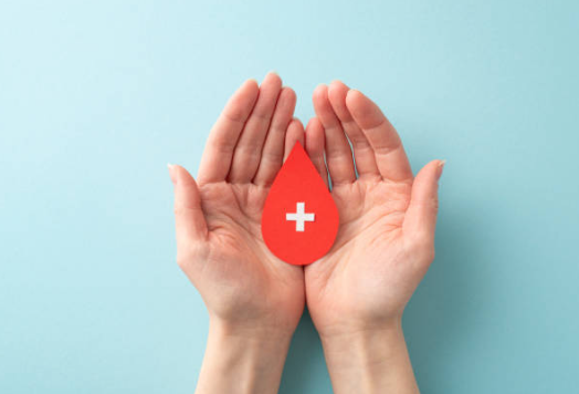 Hands cupping a red blood drop with a white cross, on a blue background.