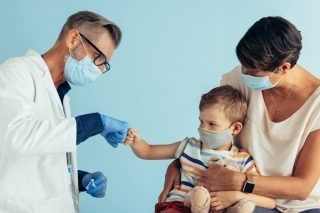 Doctor fist-bumps a child held by a woman; all wear masks. Medical setting, blue background.