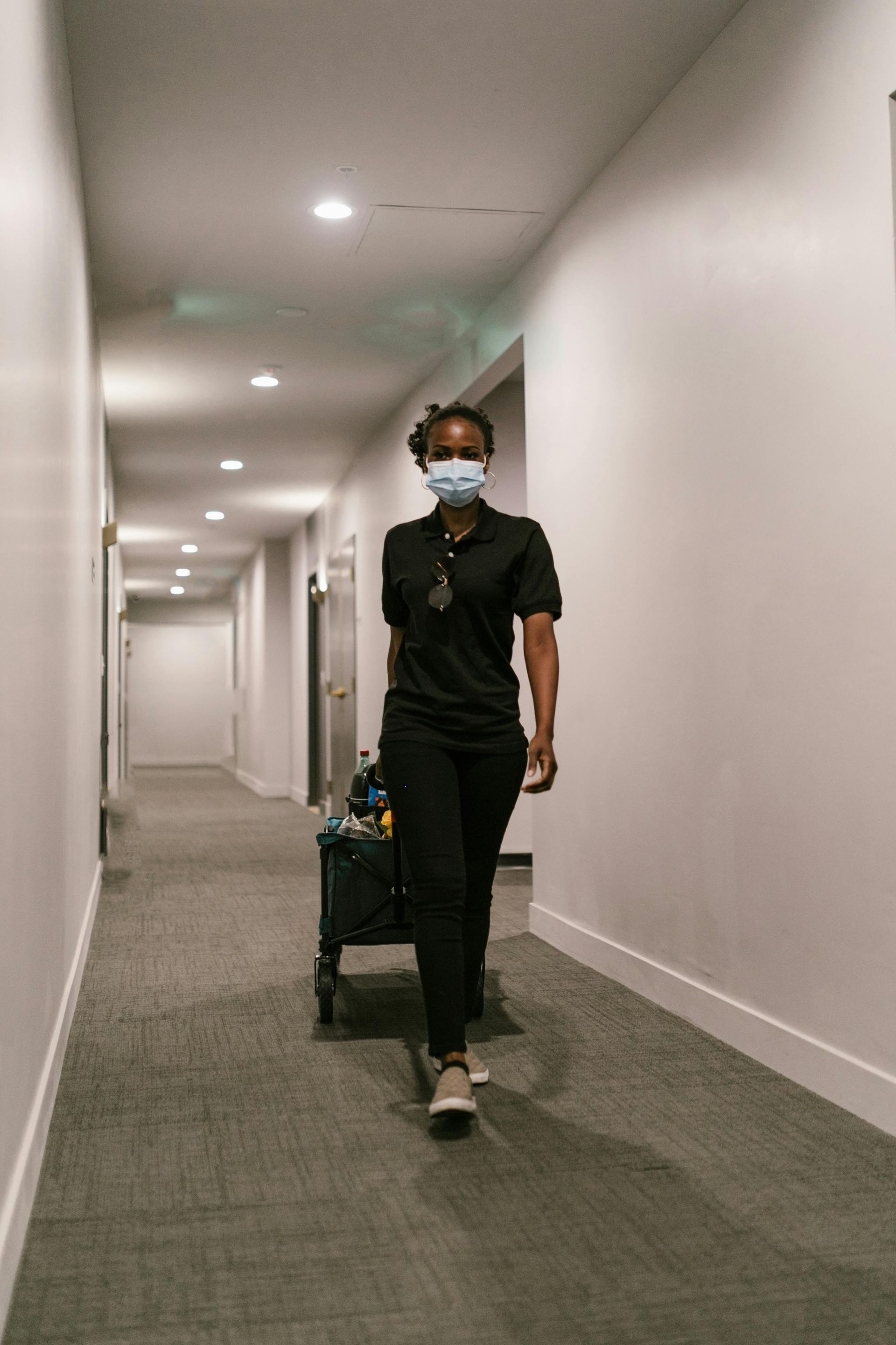 Woman in black scrubs and mask walking down a hallway, pulling a supply cart.