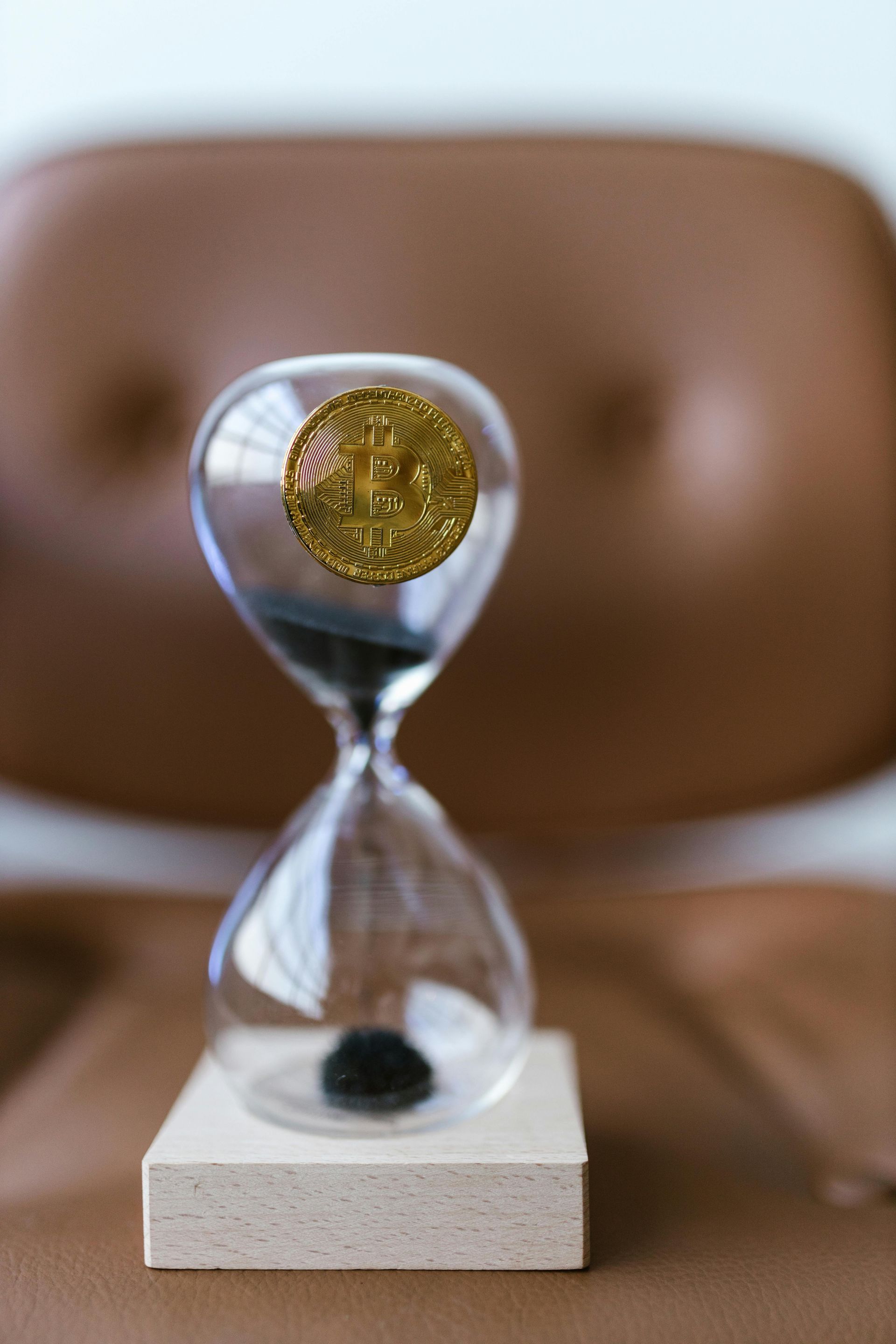 Bitcoin coin inside an hourglass, sand flowing. Set on a white block, brown chair background.