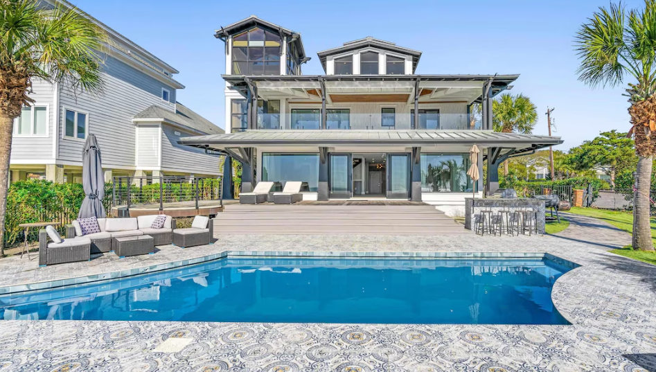 Luxury waterfront home with pool, deck, outdoor kitchen, and palm trees.