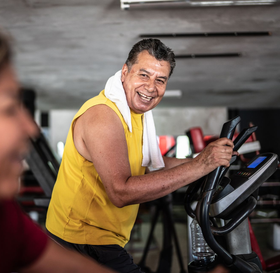 Man in yellow shirt smiles while exercising on elliptical machine at a gym.