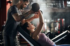 Woman doing dumbbell chest press, assisted by trainer in gym.