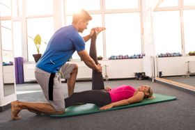 Man assisting woman with leg stretch on a mat in a gym.