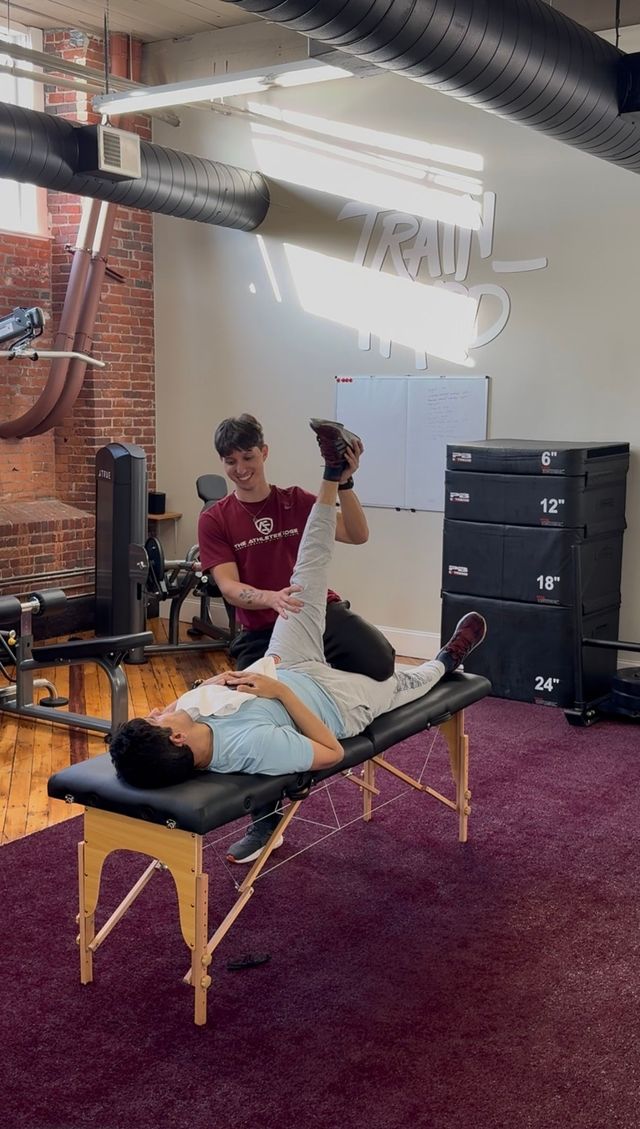 Man lying on a massage table while another man assists with leg stretch in a gym.
