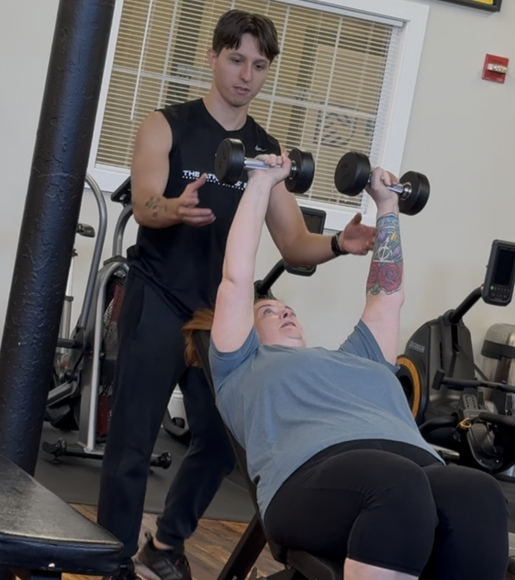 A personal trainer guides a woman doing dumbbell chest press in a gym.