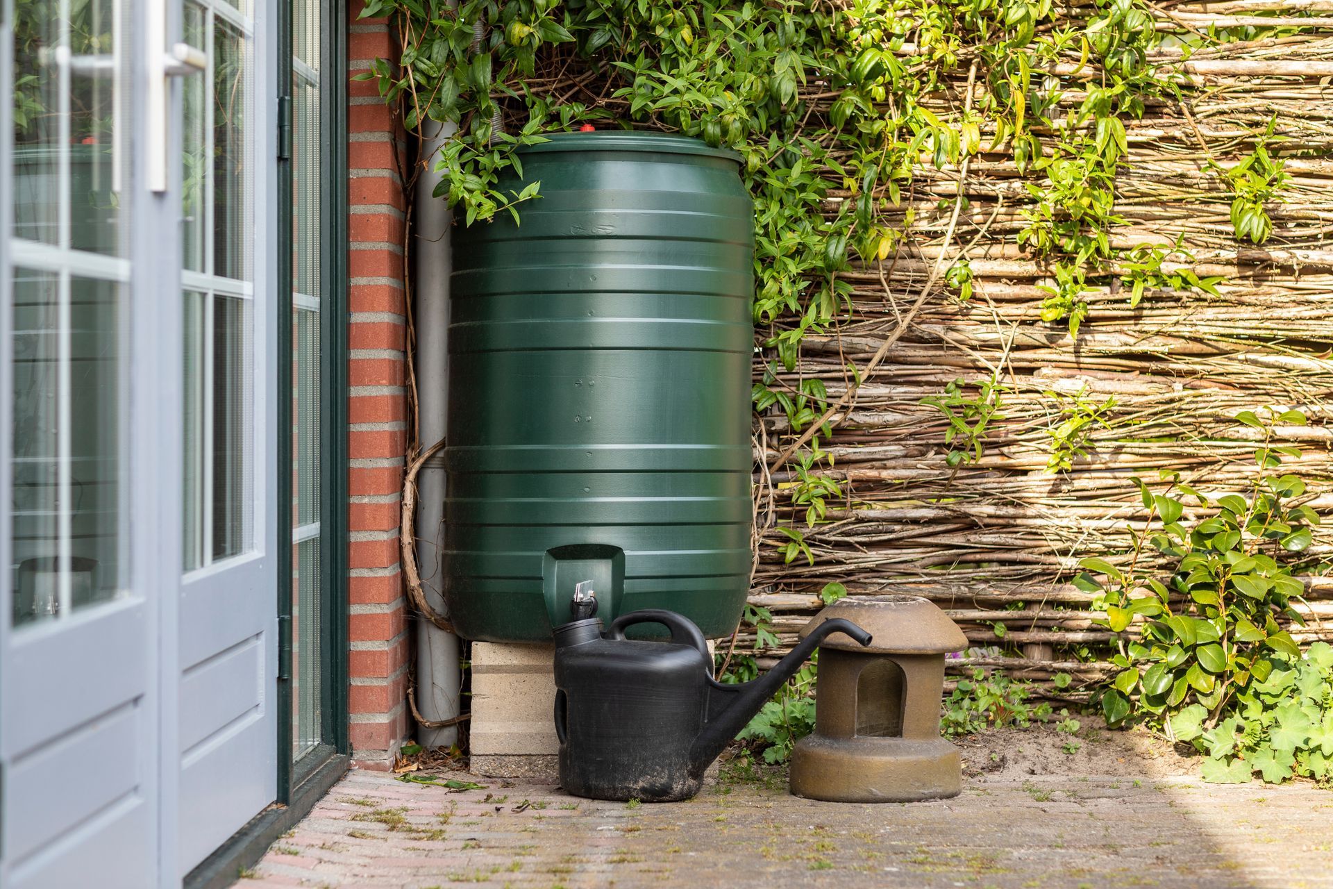 A green water tank in a backyard with a wattle fence made of willow branches on a sunny day.
