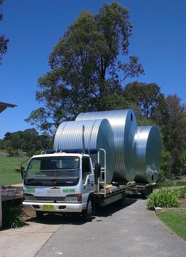 White flatbed truck carrying a large silver metal tank on a driveway with trees and blue sky behind