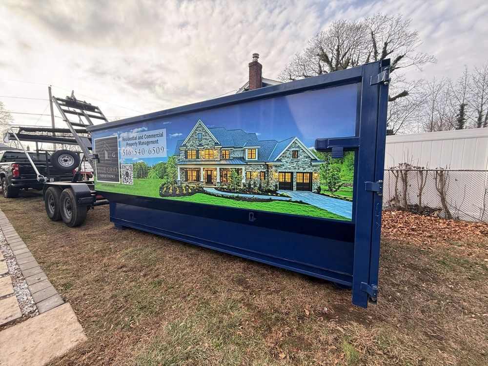 Blue dumpster trailer with a house graphic, parked on grass next to a sidewalk and truck.
